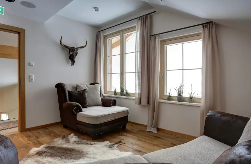 Cozy hotel room with leather armchair on sheepskin rug, longhorn skull on wall, snowy alpine views through windows.