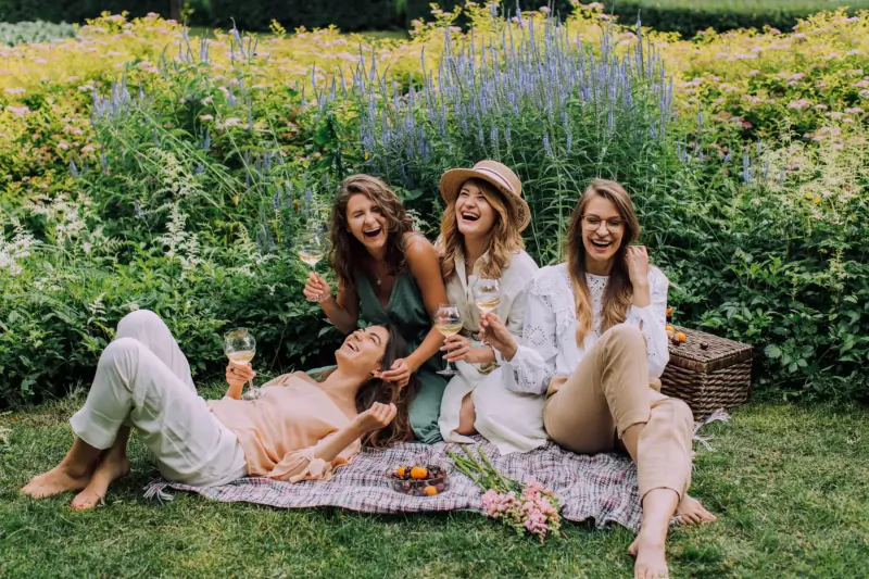 Four smiling women enjoying a posh picnic in a lush garden, holding wine glasses, with basket, flowers, and fruits on a blanket.