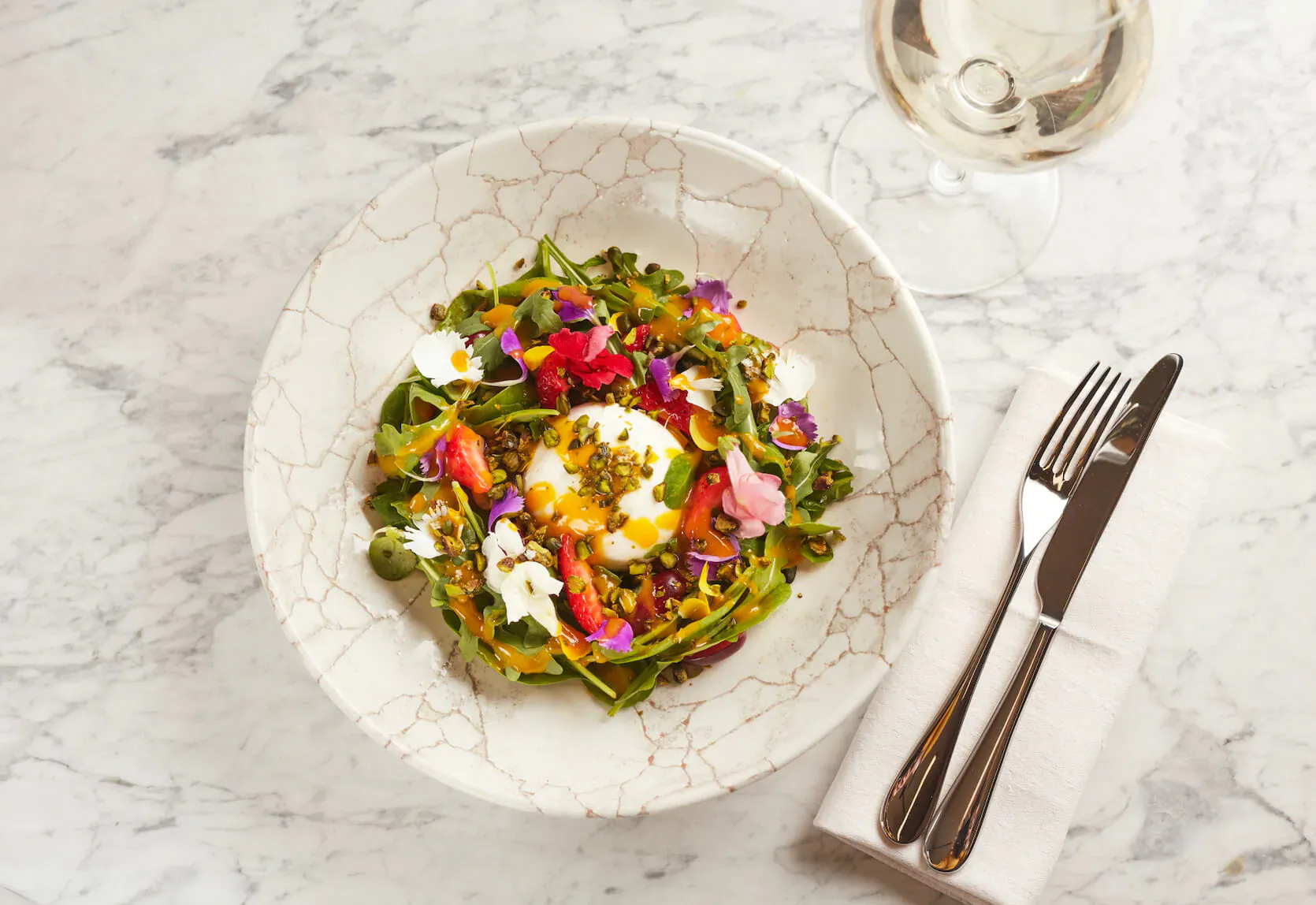 Vibrant edible flower salad with poached egg in white bowl, knife and fork, white wine glass on marble table