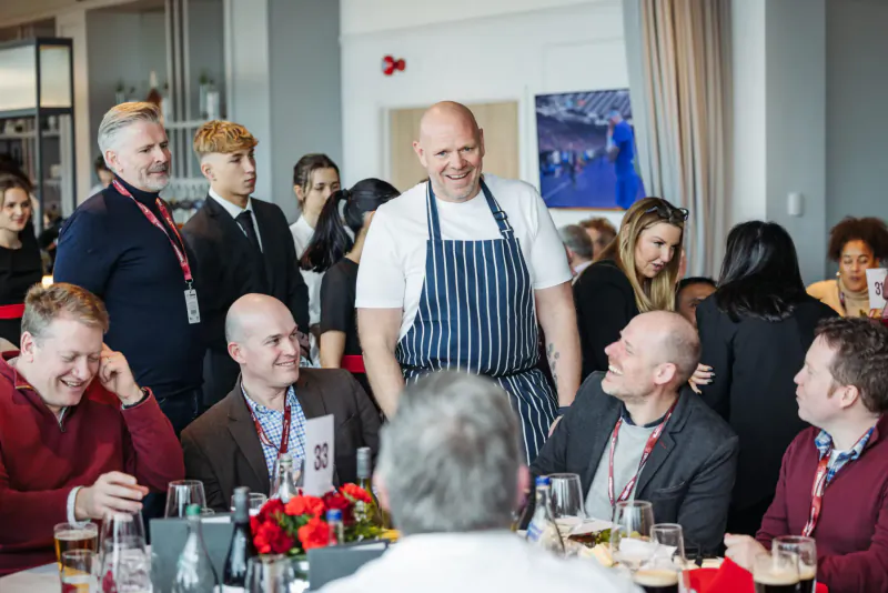 Group of smiling professionals in suits and chef in striped apron at festive holiday dinner table with wine glasses and flowers.