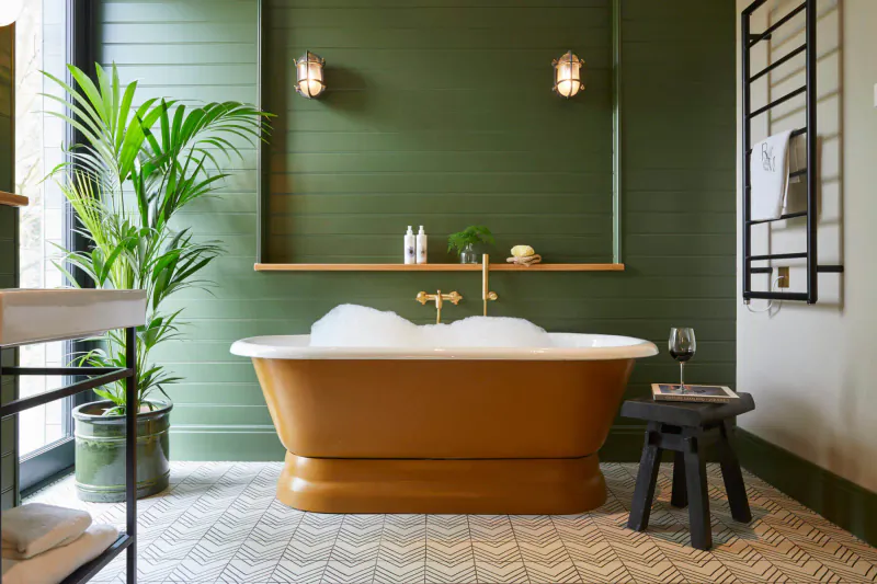Luxurious clawfoot bathtub filled with bubbles in green-tiled bathroom at Rothay Manor, with plants and wine glass.