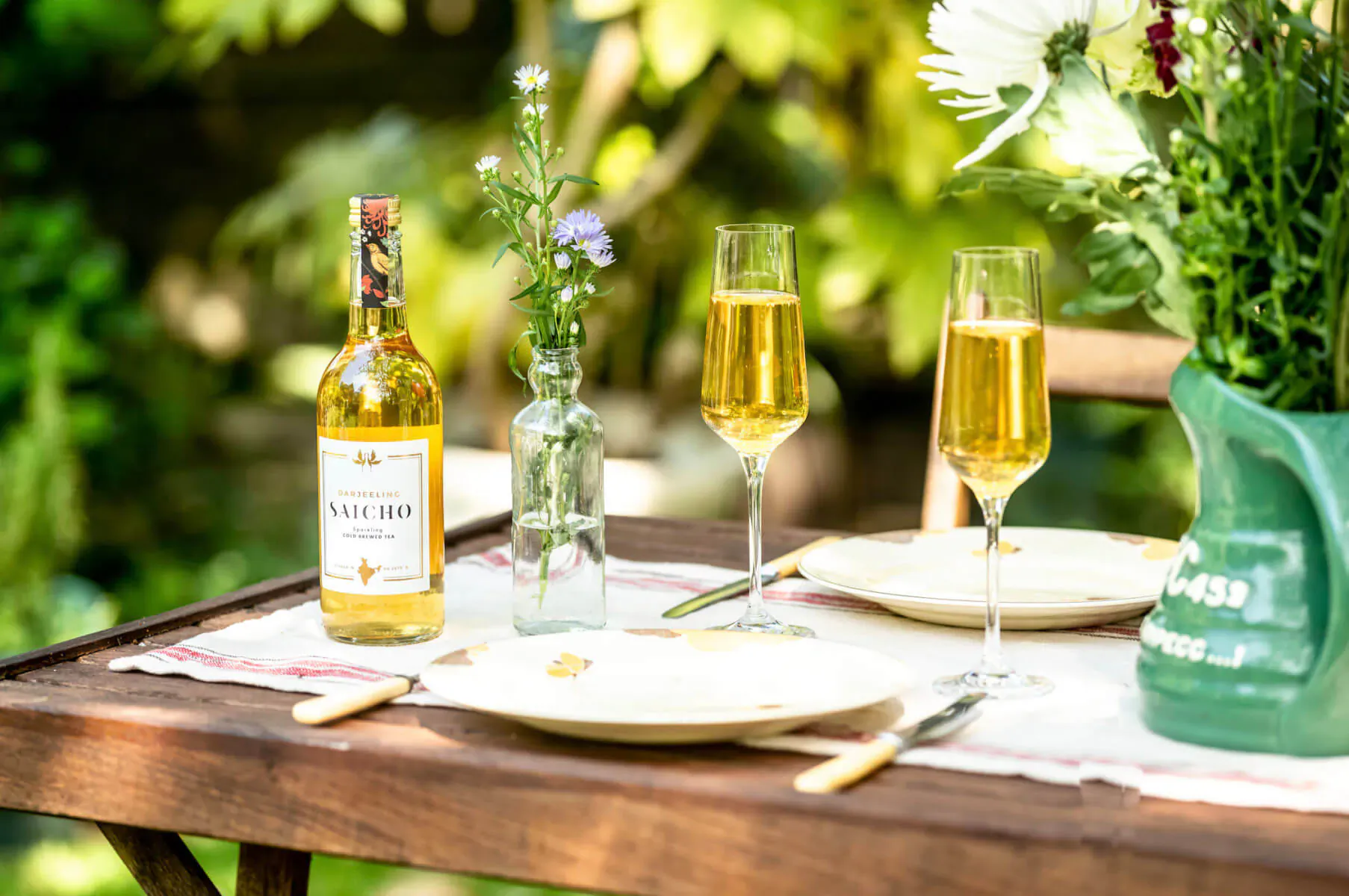 Outdoor table with Saicho sparkling tea bottle, two filled glasses, plates, cutlery, flowers, and greenery.