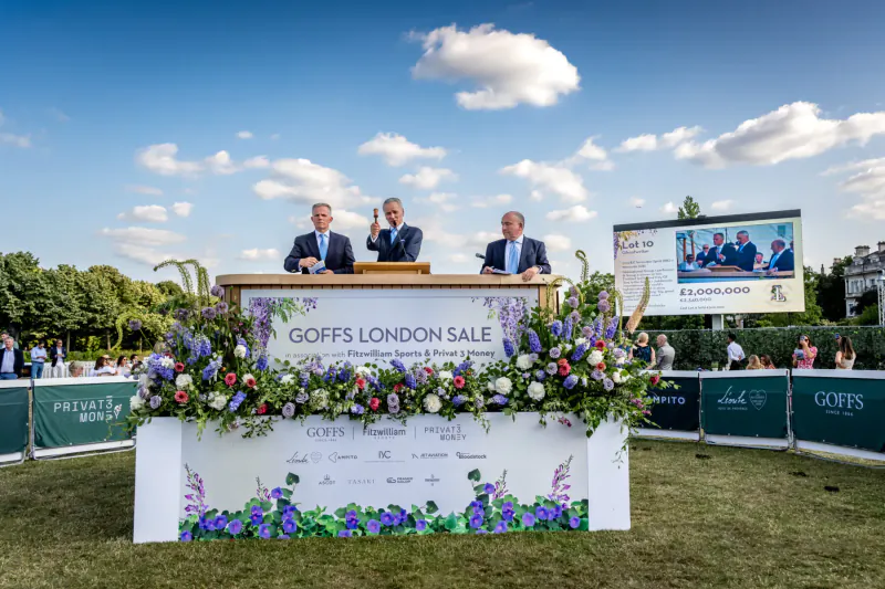 Three suited men at podium with 'Goffs London Sale' banner and flowers, outdoors under blue sky with clouds.