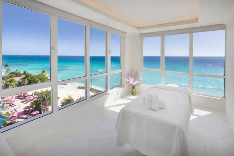Bright spa room with massage table, towels, pink flowers, and panoramic ocean views from large windows at O2 Beach Club & Spa.