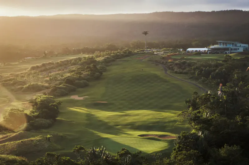 Aerial view of lush green golf course at Avalon Golf Estate, Mauritius, with clubhouse, palm trees, and misty mountains at sunset.