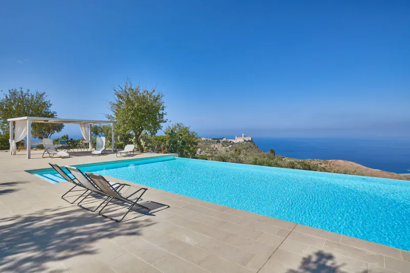 Infinity pool with loungers at luxury villa in Sicily, overlooking sea and cliffs under blue sky