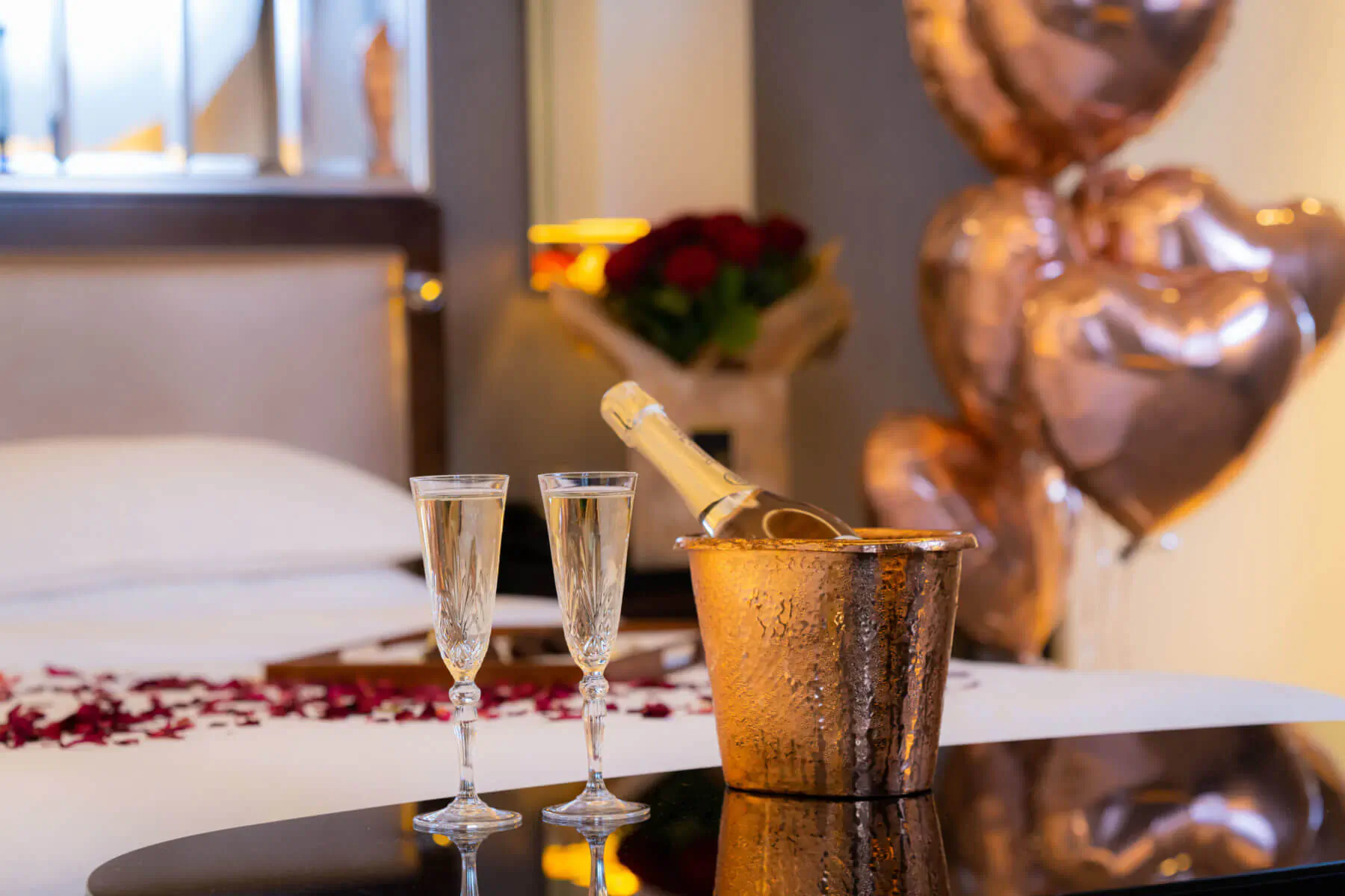 Luxury hotel bed with rose petals, two champagne flutes, bottle in copper ice bucket, red roses, and pink heart balloons for romantic celebration.