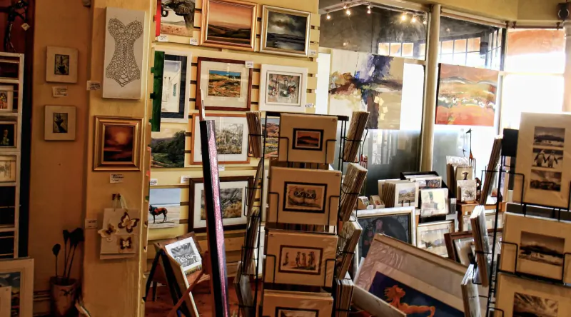 Cozy Kalk Bay art gallery with stacked frames, landscapes, and a mannequin silhouette amid warm lighting.