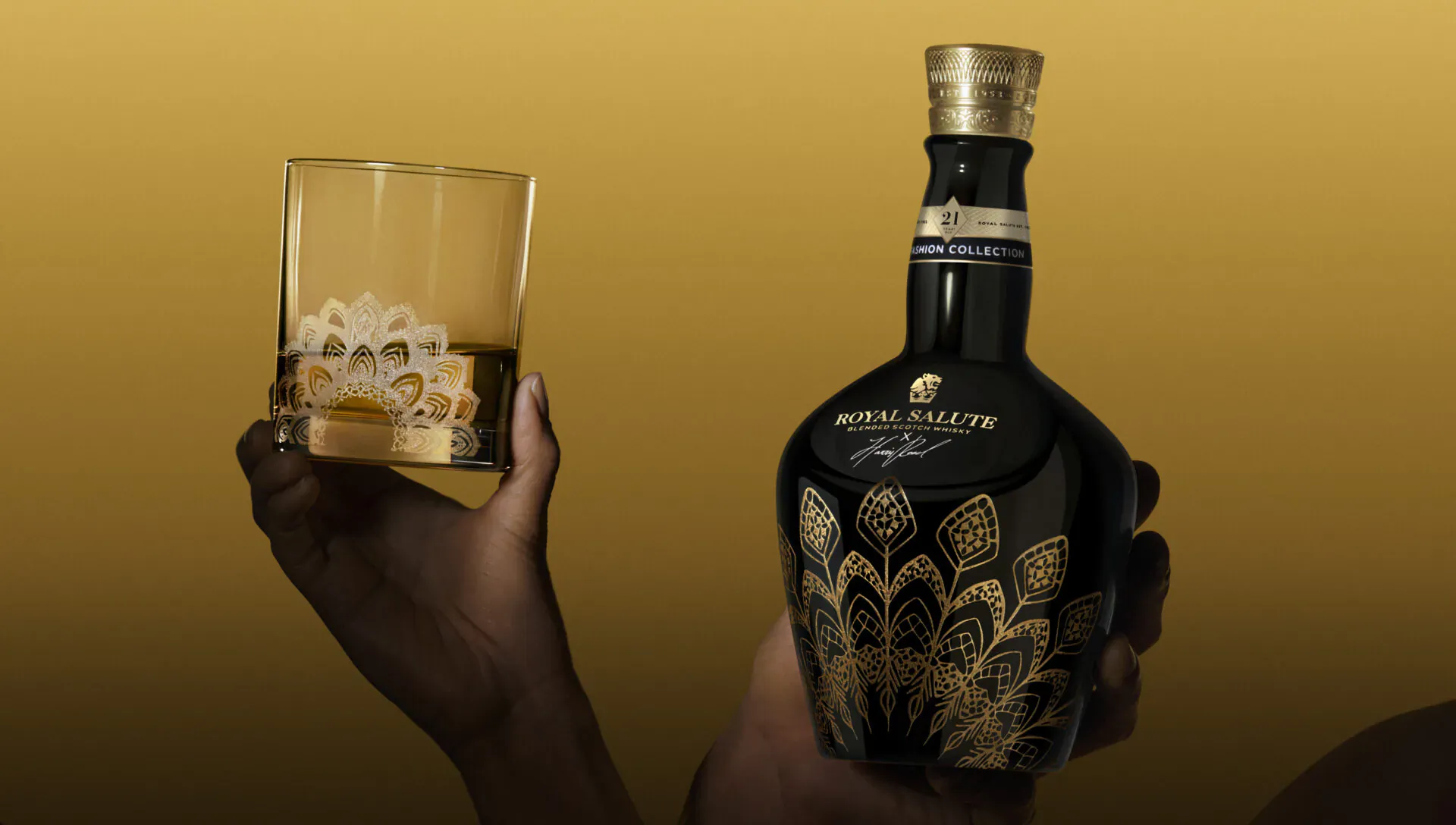 Hands holding Royal Salute whisky bottle with intricate gold peacock design and a glass of whisky on the rocks