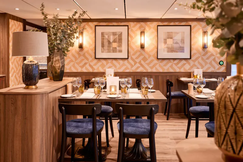 Elegant VIVA ENJOY cruise ship dining room with wooden tables, blue chairs, lamps, eucalyptus, and geometric art.