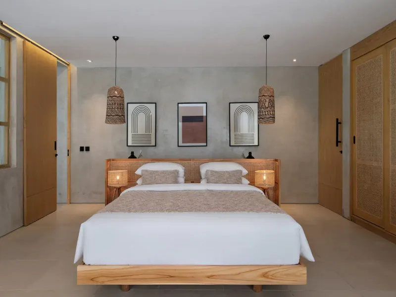 Spacious bedroom at Amber Lombok Beach Resort with wooden bed, rattan lamps, abstract art, and natural wood decor.