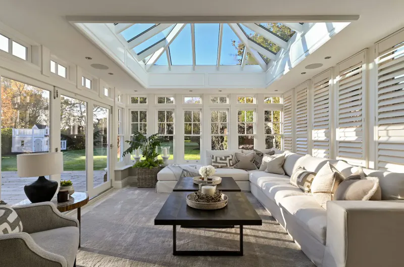 Elegant sunlit conservatory with white sofas, coffee table, plants, and garden view through glass walls.