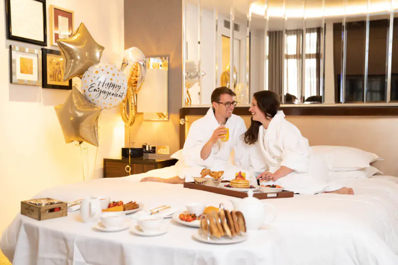 Couple in white robes on bed in Sheraton hotel room, sipping orange juice from tray with breakfast and 'Happy Birthday' gold balloons.