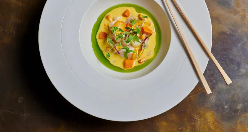 Overhead view of Nikkei dish: white plate with yellow curry sauce, orange sweet potato chunks, green onions, chopsticks on brown surface