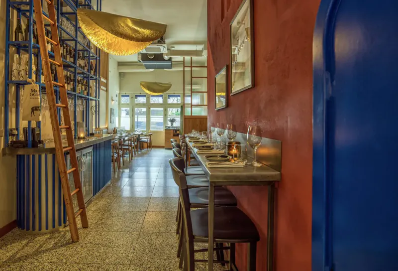 Cozy interior of Vori restaurant in London with wooden tables set for sharing, blue door, red walls, golden ceiling lamps, and bar with ladder.