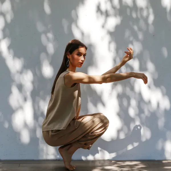 Woman in beige tank top and pants squats gracefully barefoot on sunlit floor with leaf shadows, arms extended elegantly.