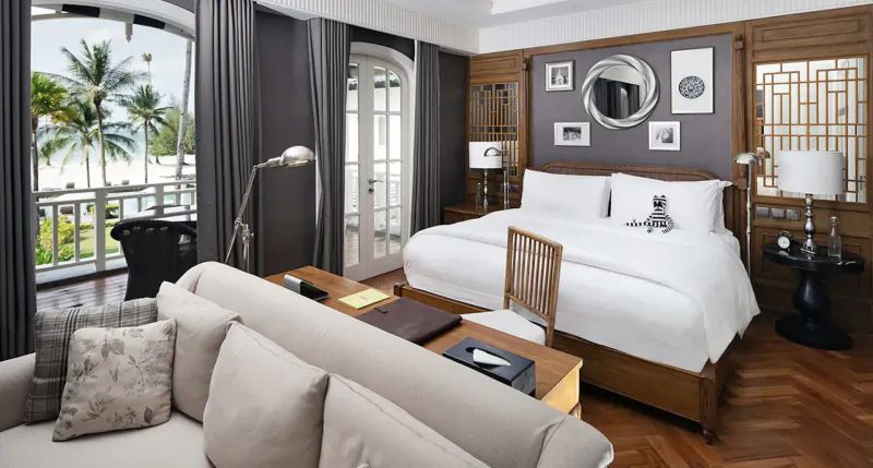 Luxurious hotel suite with king bed, plush sofa, desk, and balcony overlooking palm trees and beach.