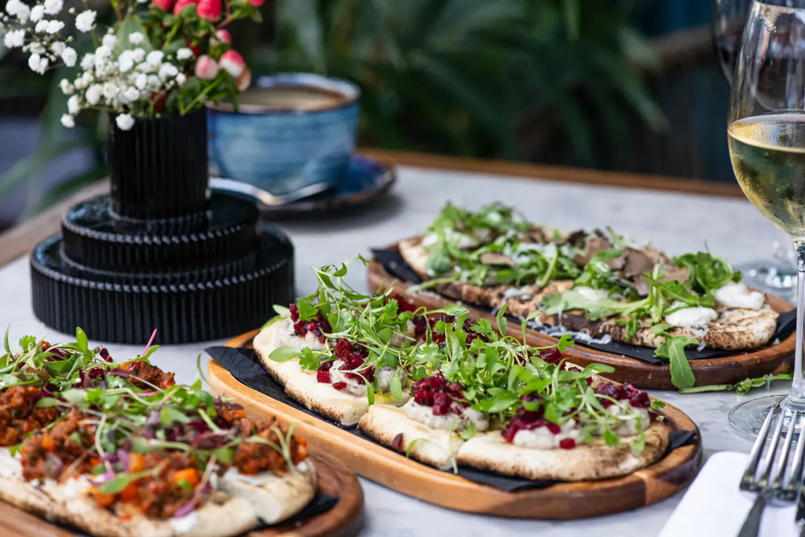 Three Mediterranean flatbreads topped with greens, feta, and pomegranate on wooden boards, with wine, coffee, and flowers on table