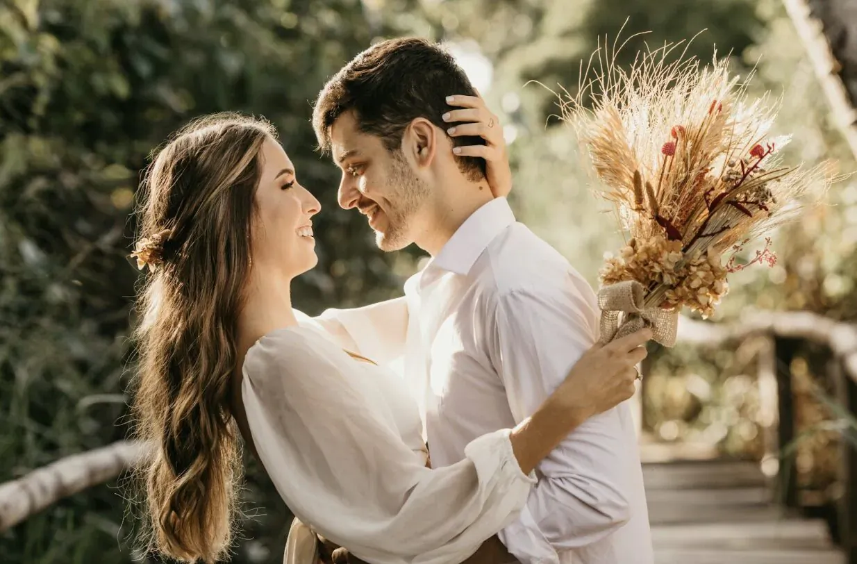 Smiling couple in white outfits embracing on wooden bridge in lush greenery, man holding dried flower bouquet