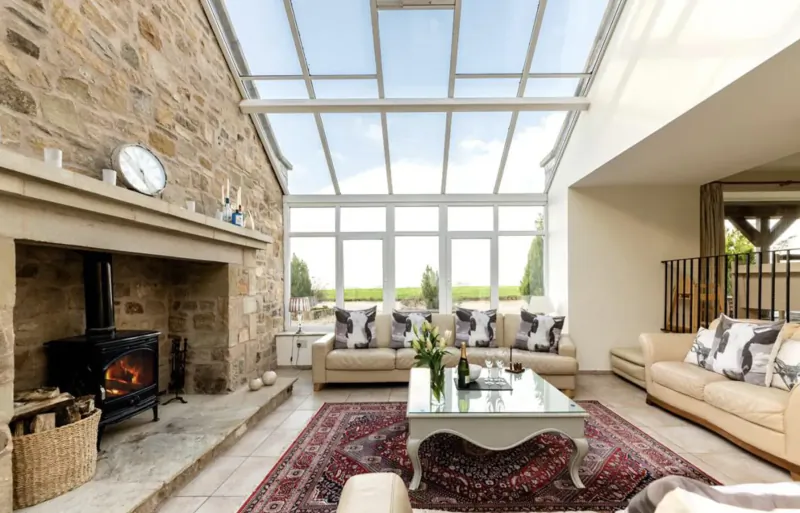 Luxury UK holiday cottage conservatory with stone walls, glass roof, lit fireplace, beige sofas, and ornate rug.
