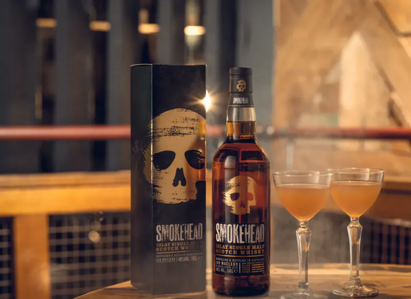 Smokehead whisky bottle and box with skull logo beside two glasses of amber whisky on wooden bar.