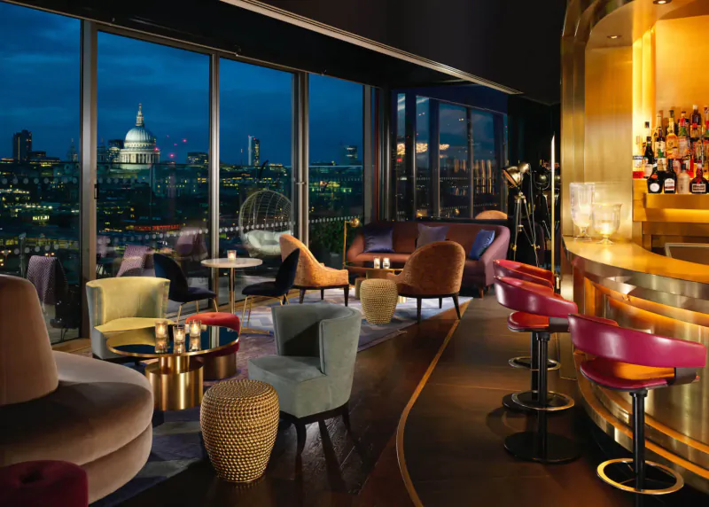Elegant spa lounge at dusk with panoramic London skyline view including St. Paul's Cathedral, plush colorful chairs, and curved gold bar.