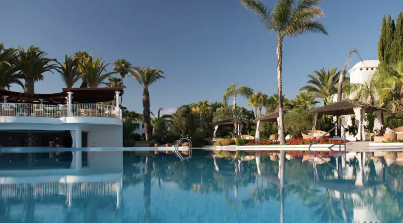 Luxury resort infinity pool with clear blue water, palm trees, white buildings, loungers, and sunny sky reflections.