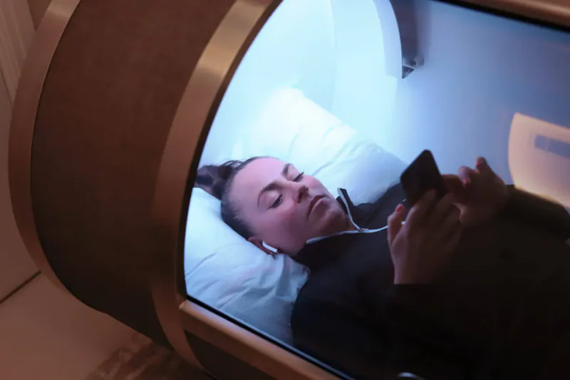 Woman lying in curved Body Lab pod, relaxed under blue light, checking phone with AirPods in.