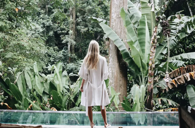 Blonde woman in white kimono stands barefoot by infinity pool overlooking lush jungle at Sungai Jungle Villas, Bali