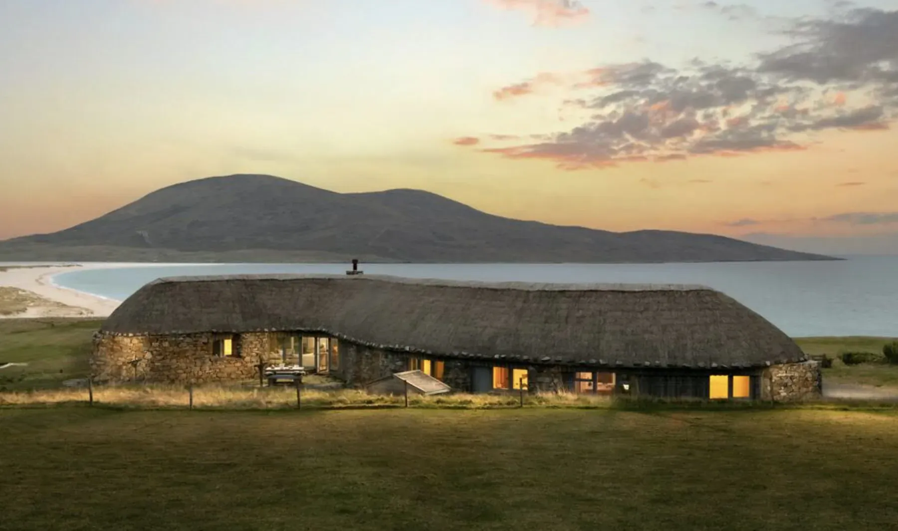 Luxury thatched cottage with stone walls on grassy coastal field at sunset, beach and mountains in background.