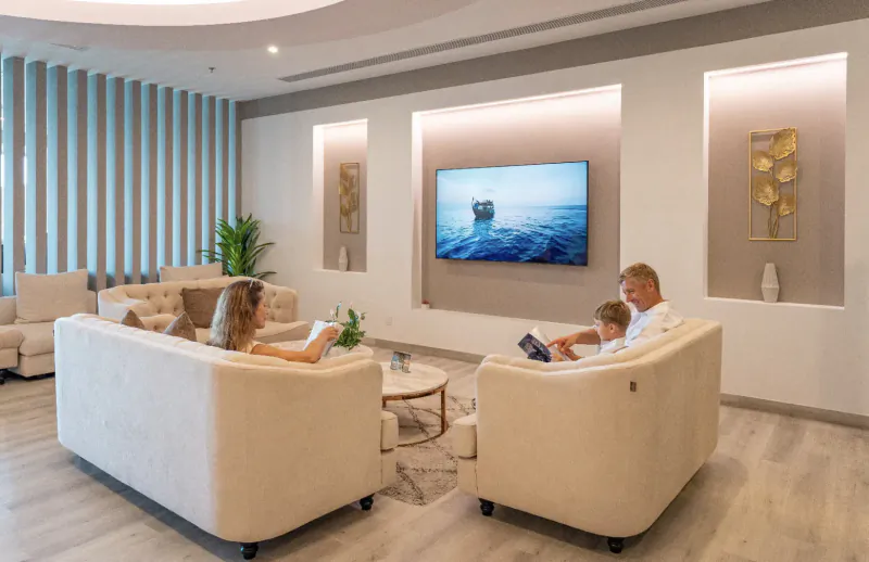 Family reading books on cream sofas in modern Lily Hotels lounge at Noovilu Seaplane Terminal, ocean TV on wall.