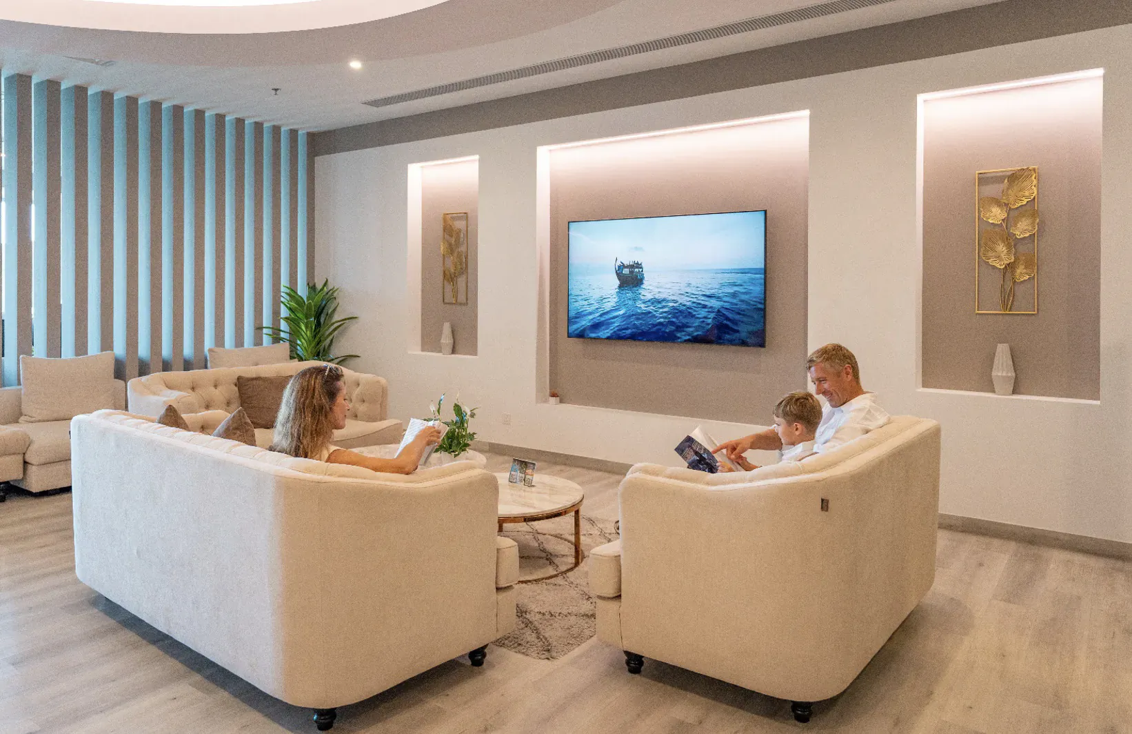 Family reading books on cream sofas in modern Lily Hotels lounge at Noovilu Seaplane Terminal, ocean TV on wall.