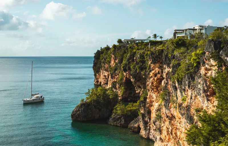 White sailboat anchored in turquoise sea by lush red cliffs with luxury resort on clifftop, blue sky.