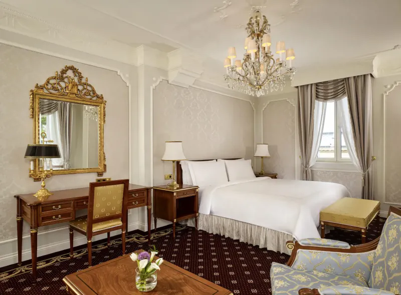 Luxurious Hotel Imperial Vienna bedroom with ornate chandelier, king bed, gilded mirror, desk, and armchair.