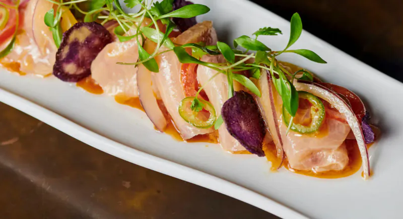 Sliced salmon sashimi with purple onions, jalapeños, tomatoes, and microgreens on white rectangular plate