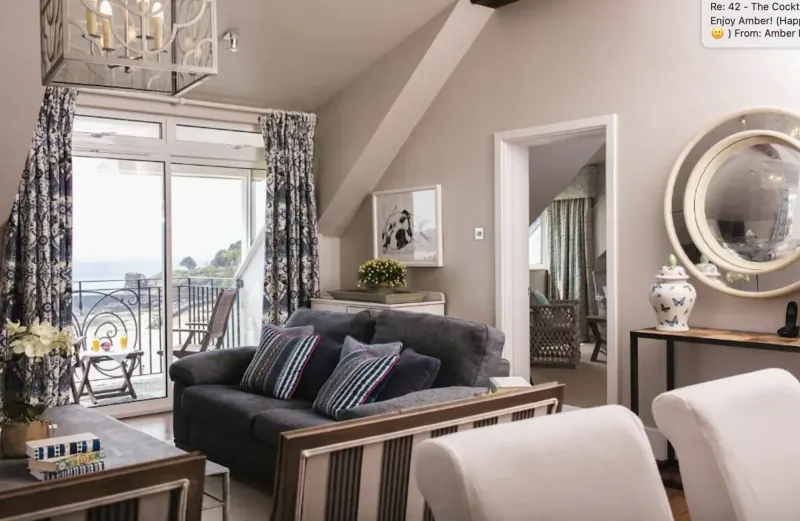 Cozy hotel living room at St. Brelade’s Bay with gray sofa, chairs, chandelier, balcony view of sea, and art.