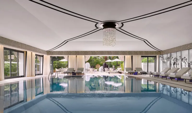 Elegant indoor pool at Hotel Metropole Monte-Carlo with lounge chairs, chandeliers, and garden views.