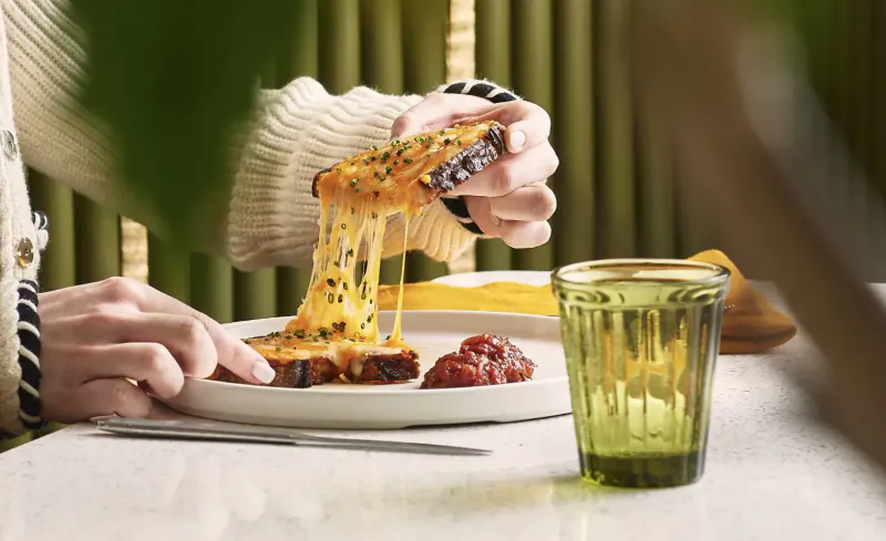 Close-up of hands pulling apart a gooey grilled cheese sandwich on a plate with green glass and ketchup at Park Corner Brasserie.