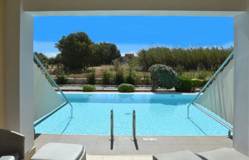 View from Cavo Spada resort lounge through shades to infinity pool, gardens, trees, and blue sky in Crete.