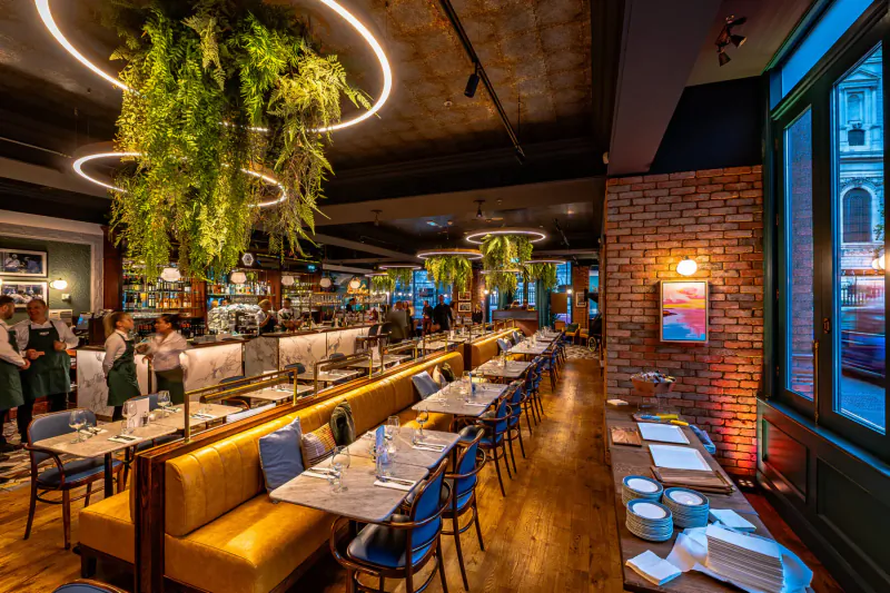 Amalfi Ristorante interior: stylish dining area with hanging plants, brick walls, yellow booths, blue chairs, staff at counter, and tables set.