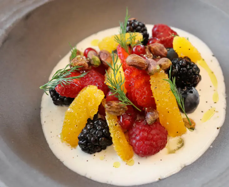 Vibrant fruit salad on creamy yogurt base with raspberries, blackberries, orange segments, currants, pistachios, and dill sprigs
