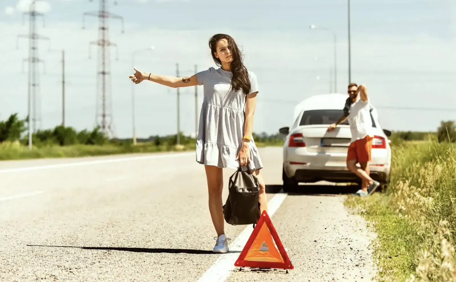 Woman in white dress hailing help with hand raised beside white car, man behind it, red warning triangle on roadside emergency.