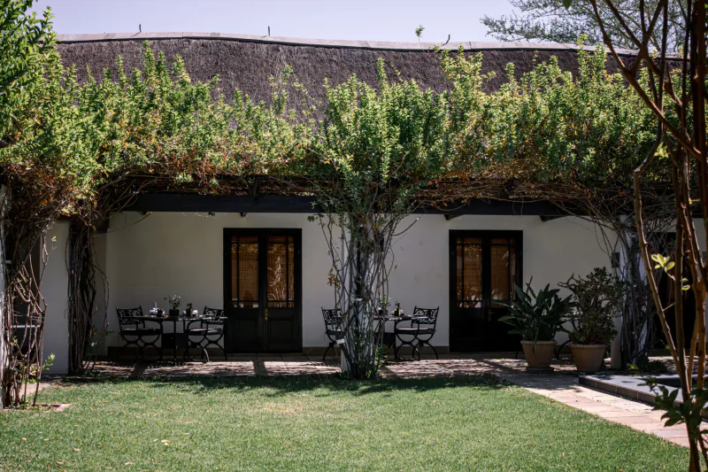 White-walled building with thatched roof and vine-covered pergola over outdoor table and chairs in lush green garden courtyard