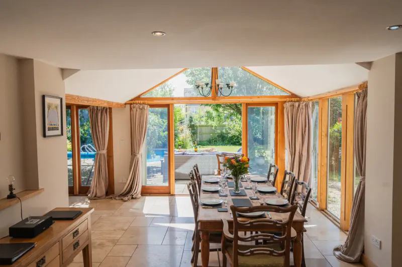 Bright Cotswolds dining room with wooden beams, long oak table set for meal, French doors to garden pool.