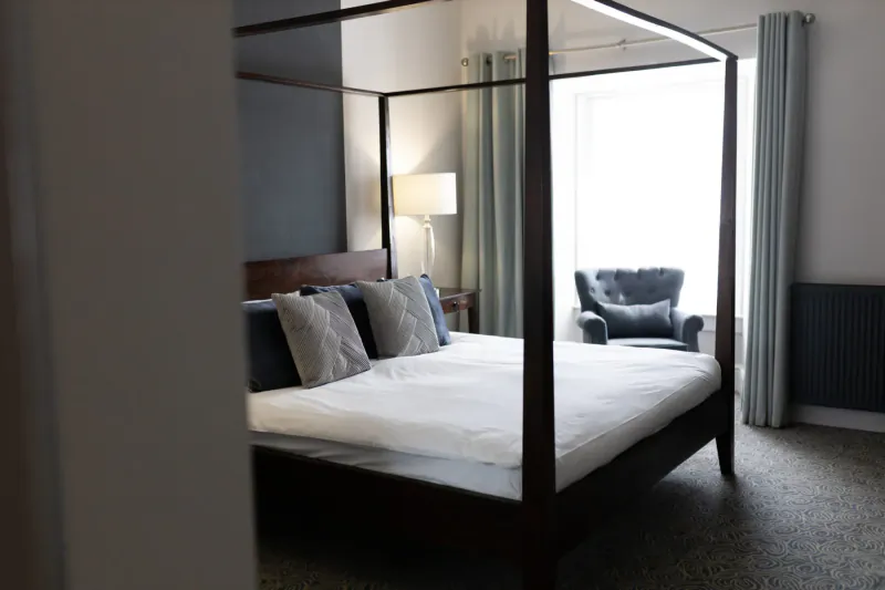 Elegant hotel bedroom with four-poster bed, blue pillows, bedside lamp, armchair, and window with curtains