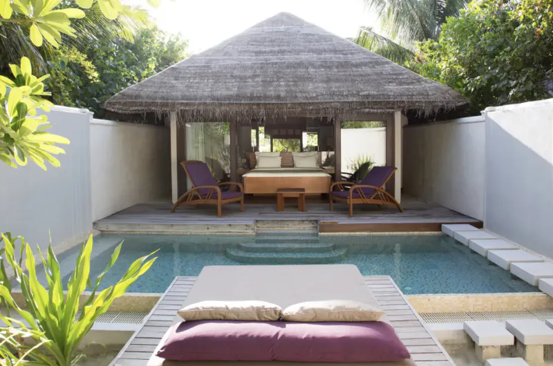 Luxurious thatched-roof overwater villa at Coco Bodu Hithi resort with private infinity pool, purple lounge chairs, and bed.