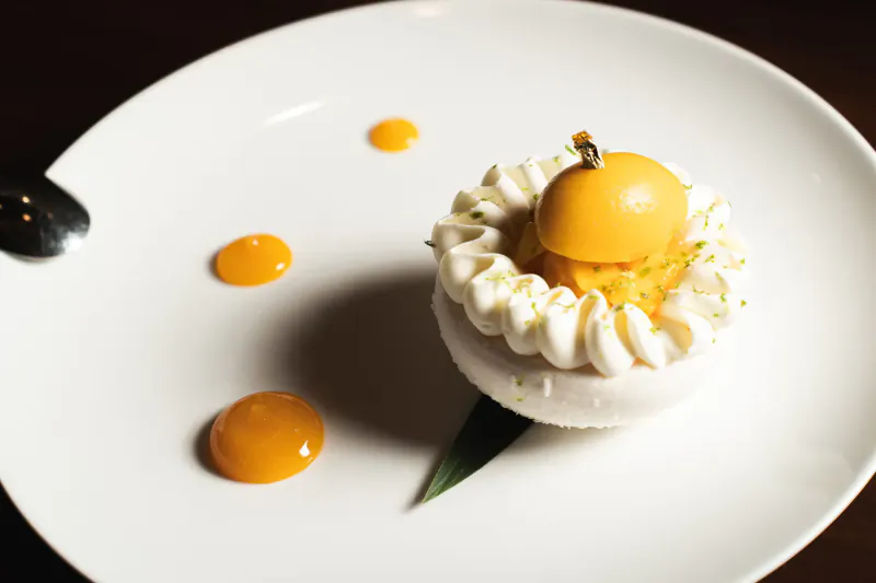 Elegant plated dessert: white meringue dome topped with gold kumquat, lime zest, orange sauce dots, shiso leaf on white plate