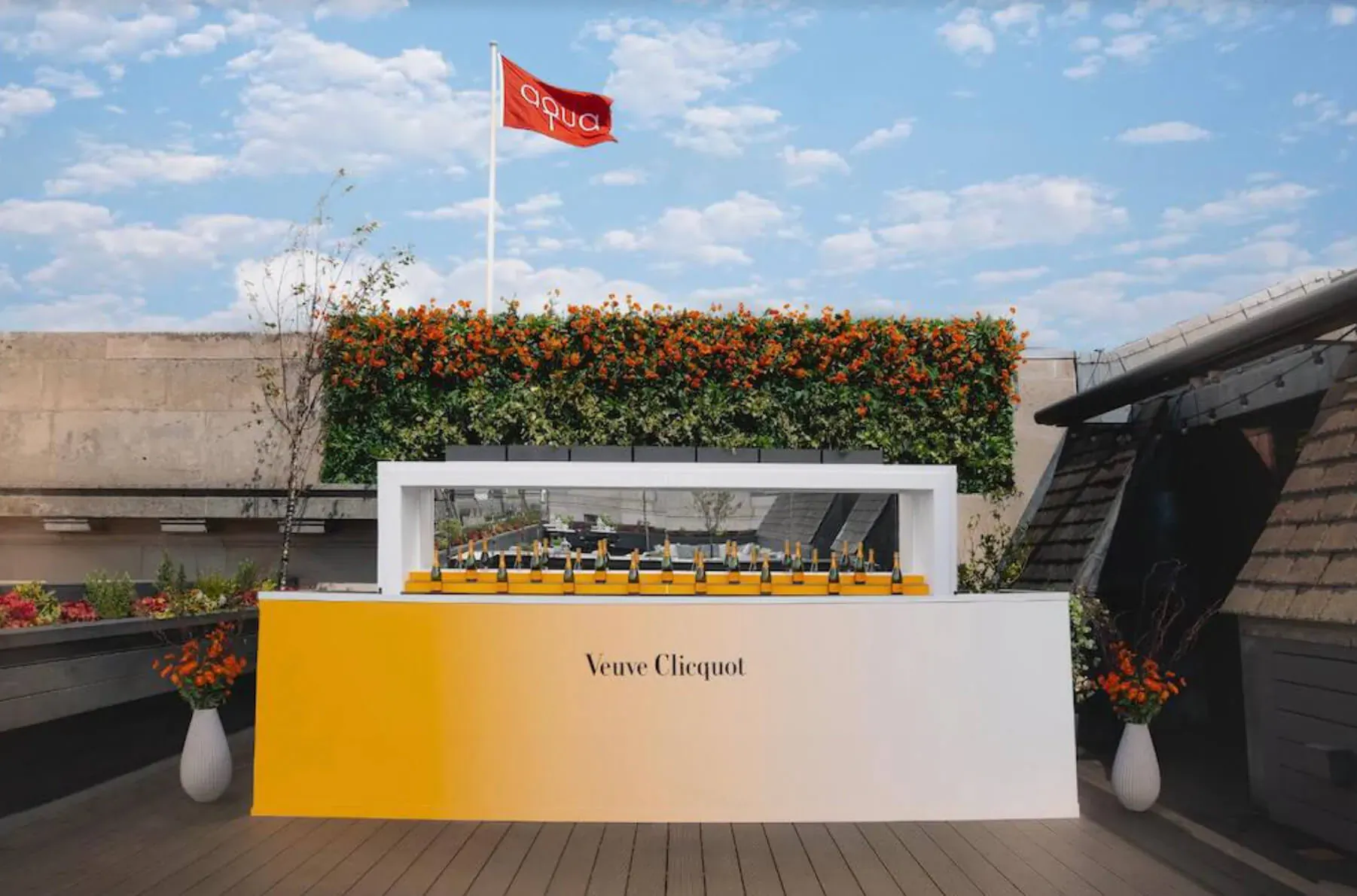 Veuve Clicquot bar on summer terrace with yellow Veuve Clicquot sign, orange flower wall, red flag, and potted plants.
