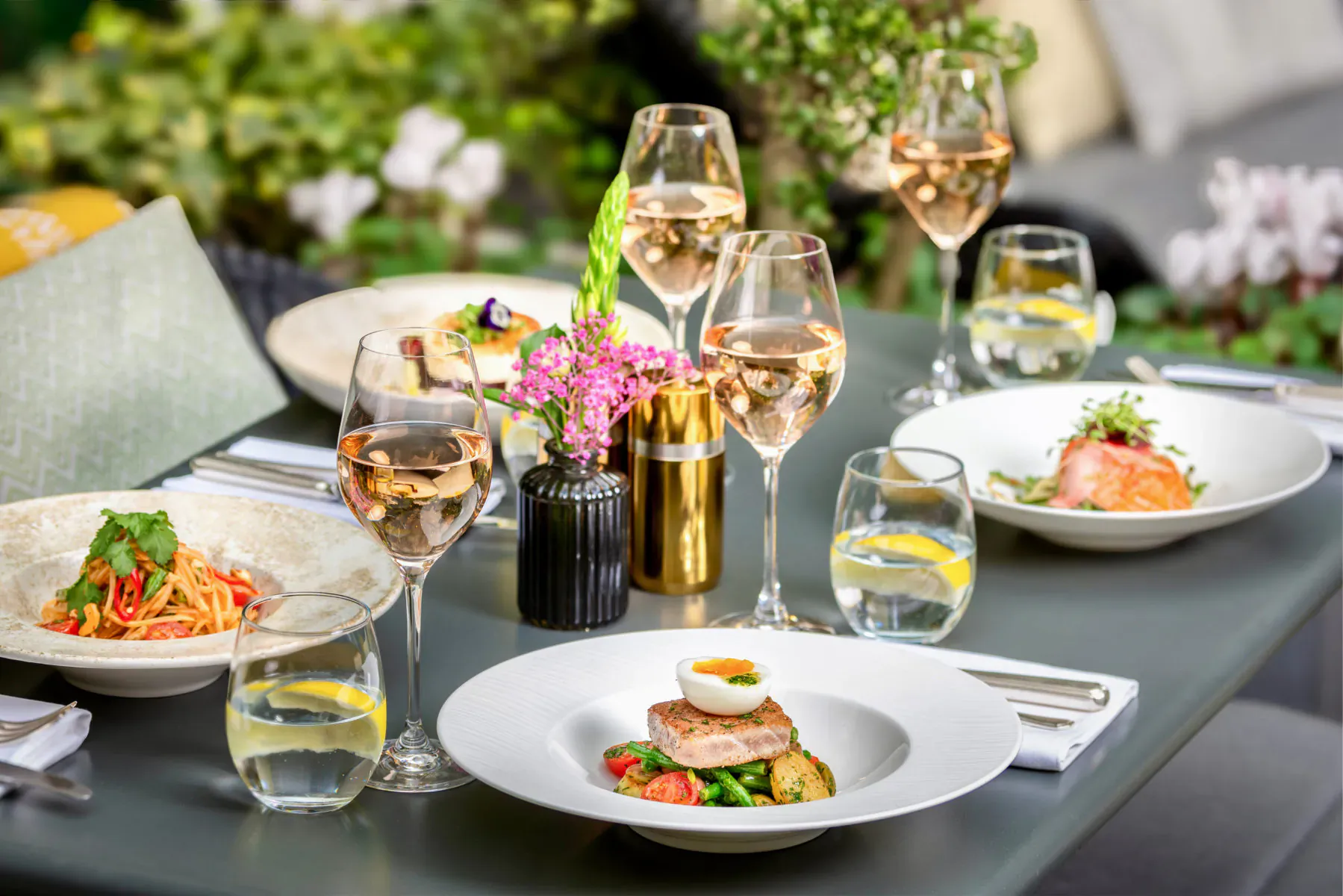 Luxurious roof terrace dining at The Sloane Club: rosé wine glasses, salmon and pasta dishes on a green outdoor table.