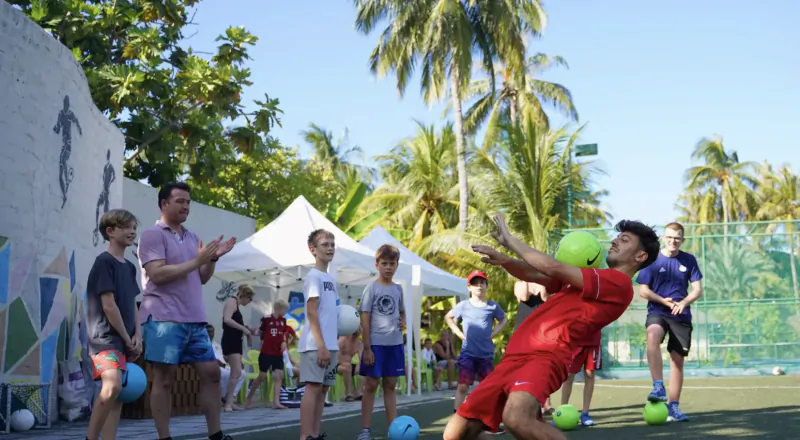 Young player in red kit kicks green soccer ball at kids' football camp on tropical resort tennis court, surrounded by cheering kids and palms.
